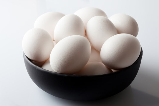 White eggs in a black bowl on white background from high angle with shadow