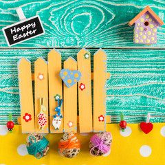 Funny bunny celebrate Happy Easter! Turquoise colored Wooden Background, yellow fence, easter eggs decorated by colorful threads, nesting box, yellow napkin at white polka dots, top view.