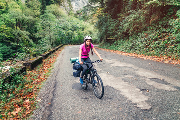 Young pretty woman with backpack and helmet travels with bicycle outdoors on the asphalt road
