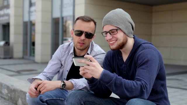 Two, handsome boys sitting near building and using cellphones. One man is showing to another something funny on his phone.
