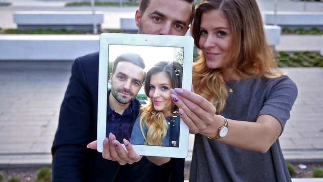 Young couple  sitting on a bench. Man is holding tablet pc and trying to take a  selfie. Positive emotions.