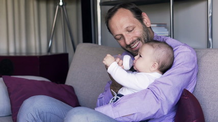 Happy father feeding his baby boy sitting on sofa at home
