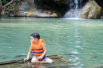 A cute boy wear colorful life jacket sitting on the wood at wate