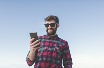 Spain, La Coruna, portrait of smiling hipster with sunglasses looking at his phablet in front of blue sky
