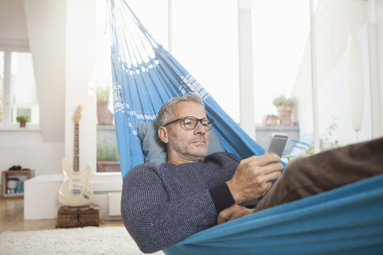 Mature man at home lying in hammock looking at cell phone