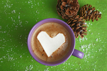 Cup of hot cappuccino with heart marshmallow and pine cones on green background, close up