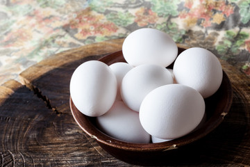 White eggs in a brown clay cracked bowl on wooden old weathered cracked cutting board on rough colourful rural map from side