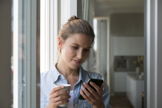 Young woman standing at the window with smart phone and coffee cup