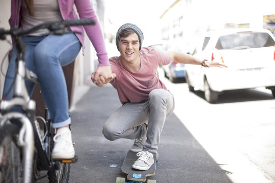 Teenage girl on bicycle pushing young man on skateboard