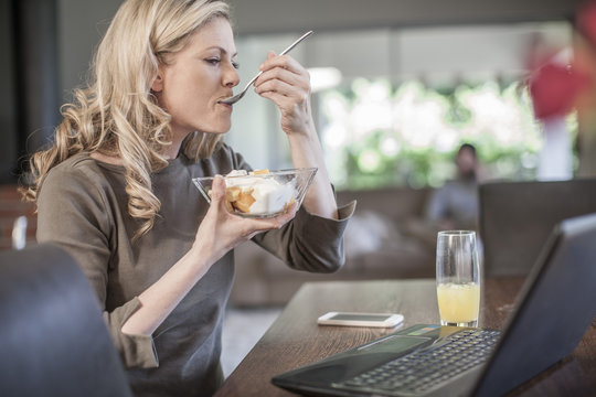 Woman eating fruit salad working at home on laptop