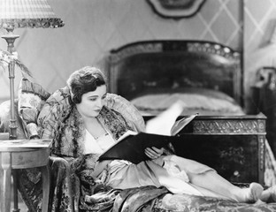 Young woman reclining in an armchair and reading a book in her bed room 