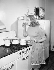 Young woman in an apron in her kitchen tasting her food from a pot 