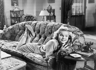 Young woman lying on a sofa in a living room and talking at the phone 