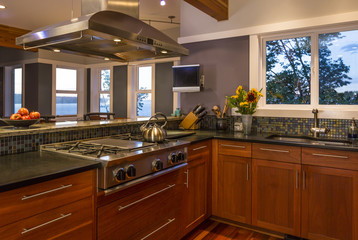 Fototapeta premium Contemporary upscale home kitchen interior with wood cabinets, gas stove, stainless steel vent hood, concrete countertops, glass tile backsplash, flat screen television and view windows