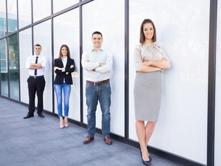 Four young and successful business people standing in front of the modern office building. Focus on confident young businesswoman standing with her arms crossed in front of her business team.