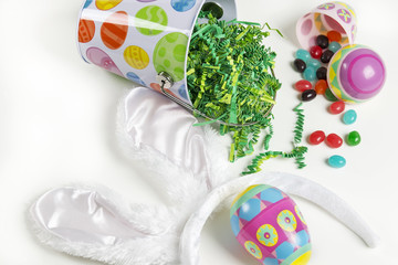 A small decorated Easter bucket with fake grass on a white background with plastic Easter eggs with spilled jelly bean candies.  A pair of white Easter bunny ears on a headband completes the scene.