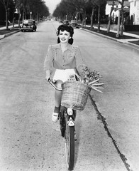 Młoda kobieta jedzie jej bicykl z koszem pełno kwiaty i marchewki - 104444405