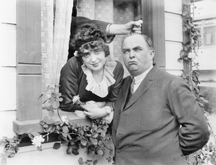 Man having his hair pulled by a hand coming out of a window while standing next to a woman 