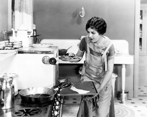 Young woman in an apron in her kitchen taking food out of the oven 