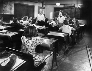 Children in a class room with a teacher and two boys looking at each other 
