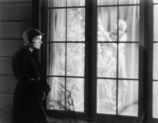 Woman standing outside of a window watching a woman trimming a Christmas tree 