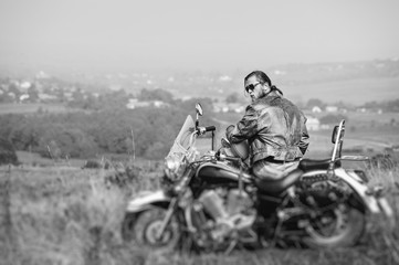 Brutal biker with beard wearing leather jacket and sunglasses sitting on his motorcycle on a sunny day, holding helmet. Horizontal picture. Rear view. Tilt shift lens blur effect. Black and white