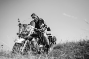 Portrait of a young biker with beard sitting on his cruiser motorcycle and looking to his bike. Man is wearing leather jacket and blue jeans. Low point of view. Tilt lens blur effect. Black and white