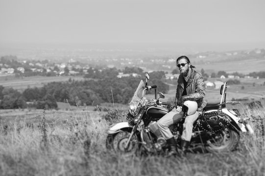 Brutal biker with beard wearing leather jacket and sunglasses sitting on his motorcycle on a sunny day, holding helmet. Horizontal picture. Tilt shift lens blur effect. Black and white