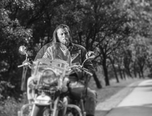Fototapeta na wymiar Portrait of biker with long hair and beard in a leather jacket and sunglasses sitting on his motobike beside the road. Looking into the camera. Tilt shift lens blur effect. black and white