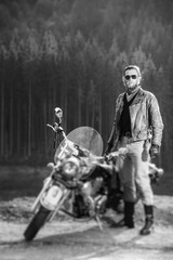 Handsome biker standing by his cruiser motorcycle on a sunny day with forest on the background. Man...