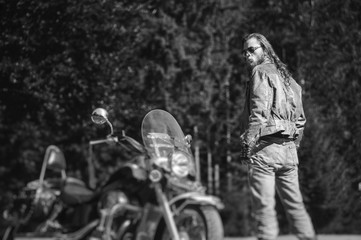 Fototapeta na wymiar Handsome brutal man with long hair and beard standing near motorcycle. Biker is wearing leather jacket and sunglasses on sunny day. Back view. Tilt shift soft effect. Black and white