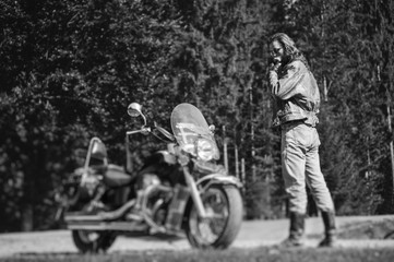 Fototapeta na wymiar Handsome biker with long hair and beard standing near his custom made cruiser motorcycle looking down. Biker is wearing jacket and sunglasses on sunny day. tilt shift soft effect. black and white