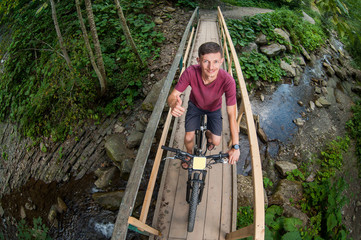 Smiling man riding his bike on the bridge crossing the mountain river and giving the thumb up. High...