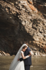 Handsome romantic groom and beautiful bride posing near river in scenic mountains