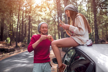 Young and lovely couple in sweaters and hats having fun, eating baguette with jam near the car on...