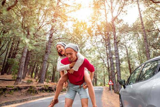 Playful young couple in sweaters and hats having a piggyback ride near the car on the pine forest roadside. Wide angle image with copy space