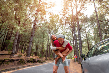 Playful young couple in sweaters and hats having a piggyback ride near the car on the pine forest...