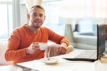 Handsome young man working on notebook, smiling, looking at camera, while enjoying coffee in cafe
