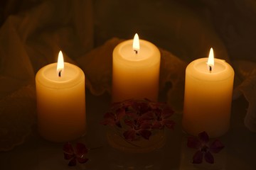 Three burning candles in darkness with a group of flowers in water, lace tablecloth in the background