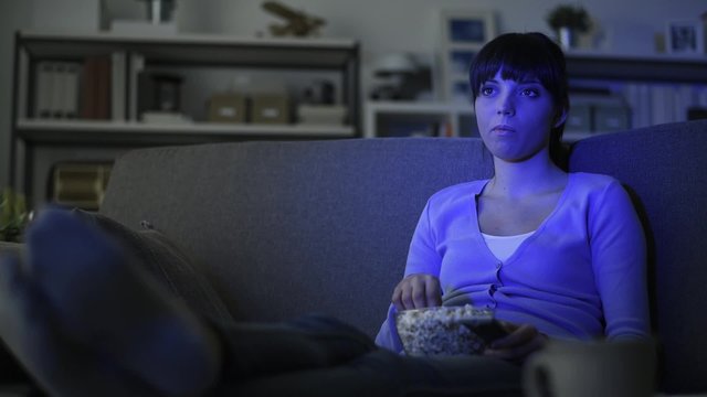 Young smiling woman relaxing on the sofa at home, she is watching tv and eating popcorn
