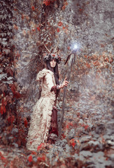 beautiful brunette with painted face, clothes shaman, a floral wreath on her head and horns, holding a glowing wooden staff, in the woods