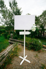 Blank wedding white banner with arrow sign decorated by flowers on stand outdoor. Copy space for your text. 