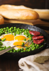 Fried eggs with sausage and green peas on a black frying pan on a wooden background