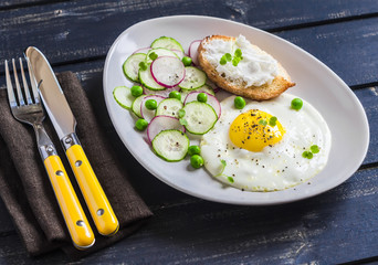 Fried egg, salad with cucumbers, radishes and green peas, toast with feta cheese on a light ceramic plate on dark wooden background. Healthy breakfast