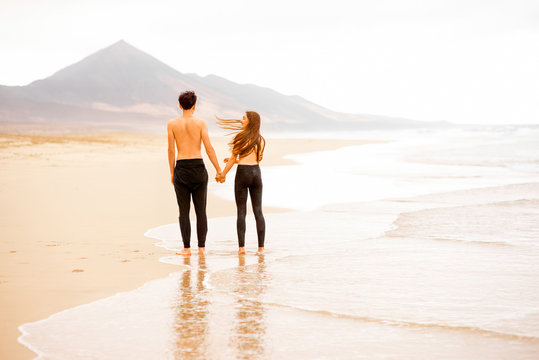 Young couple with naked torso standing and holding hands on the beautiful sandy beach with mountains on background on Fuerteventura island in Spain. General plan, rear view with copy space