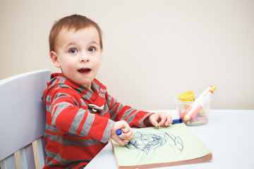 Portrait of cute Caucasian white little boy toddler drawing with color pencils markers on paper in album, looking surprised, excited, looking in camera