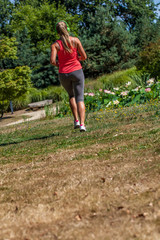 outdoors sports - back view of 20s blond woman practicing fitness with green headphones on, listening to music in park, sunny day, wide angle view