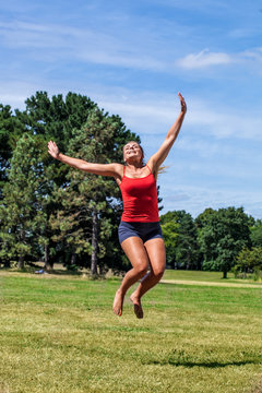 energy and fun outdoors - cheerful young blond woman laughing and jumping for success and happiness in green park, sunny summertime