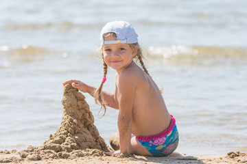 Five-year girl in a cap sits on the sand on the beach and building sand castle