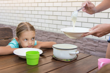 Upset six-year girl dissatisfied looks at his mother who puts porridge for breakfast